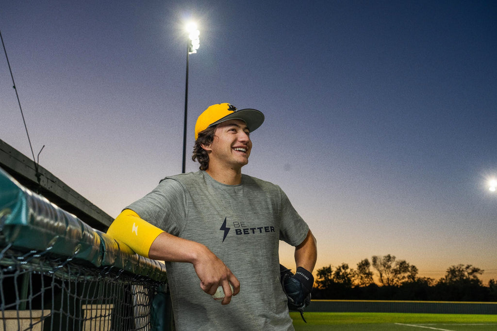 A young athlete on the side of a playing field  looking happily in the distance, wearing a BRUCE BOLT Be Better shirt