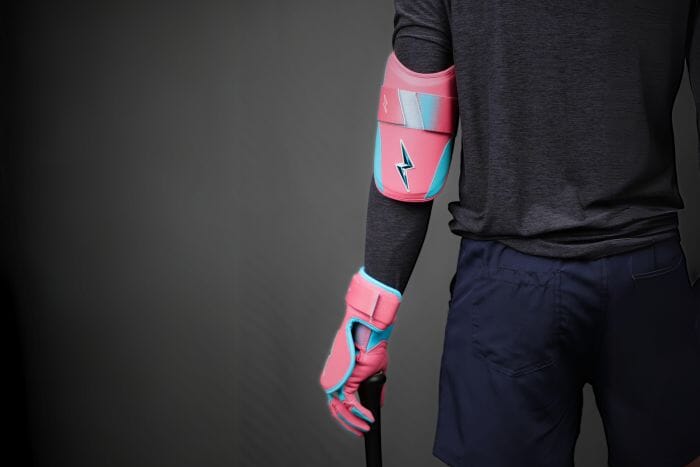 A person is facing away from the camera, wearing a pink and blue elbow and hand guard, along with a batting glove, while resting their hand on a baseball bat.