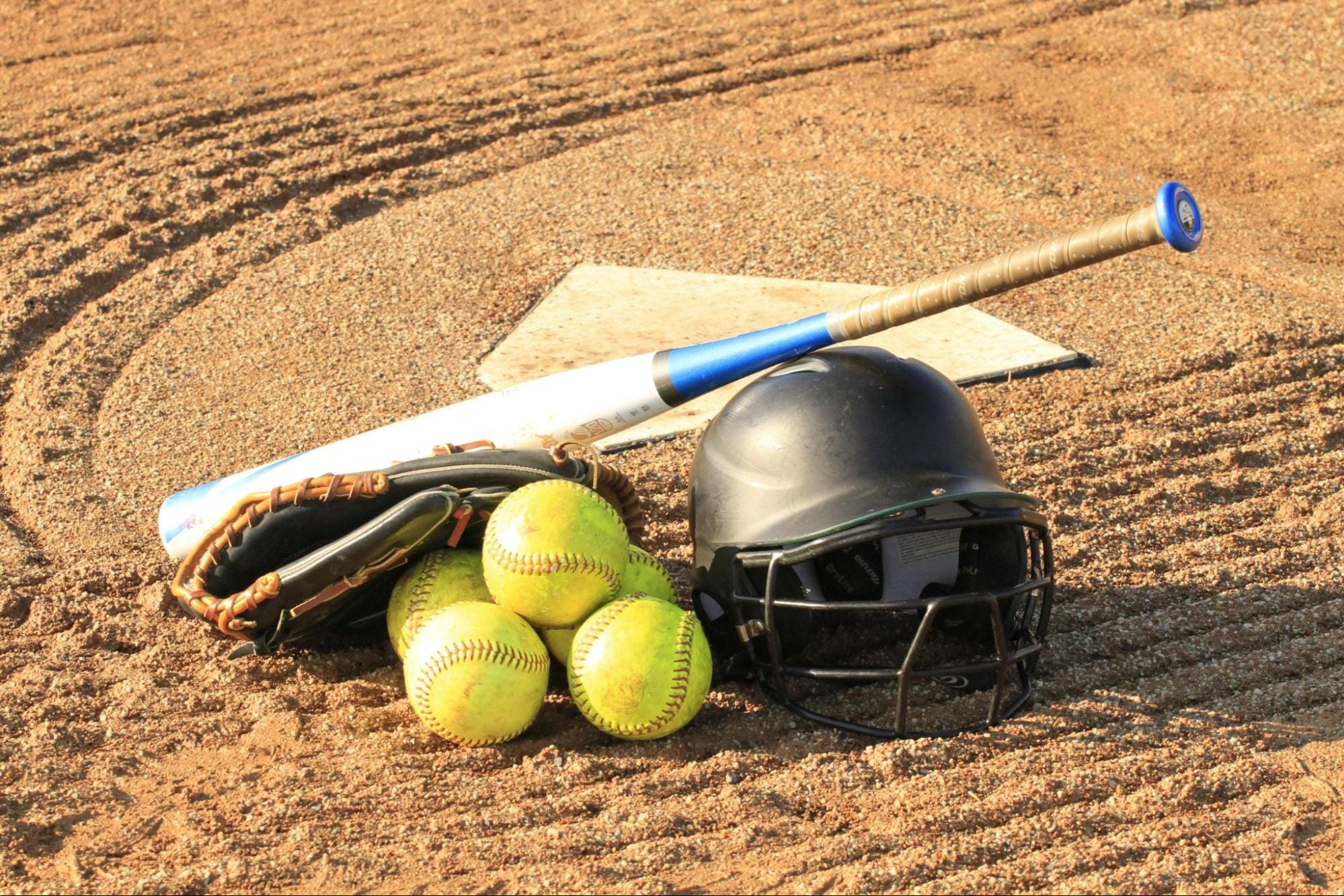 A baseball helmet, bat, catching mitt, and several baseballs are sitting on a baseball field.