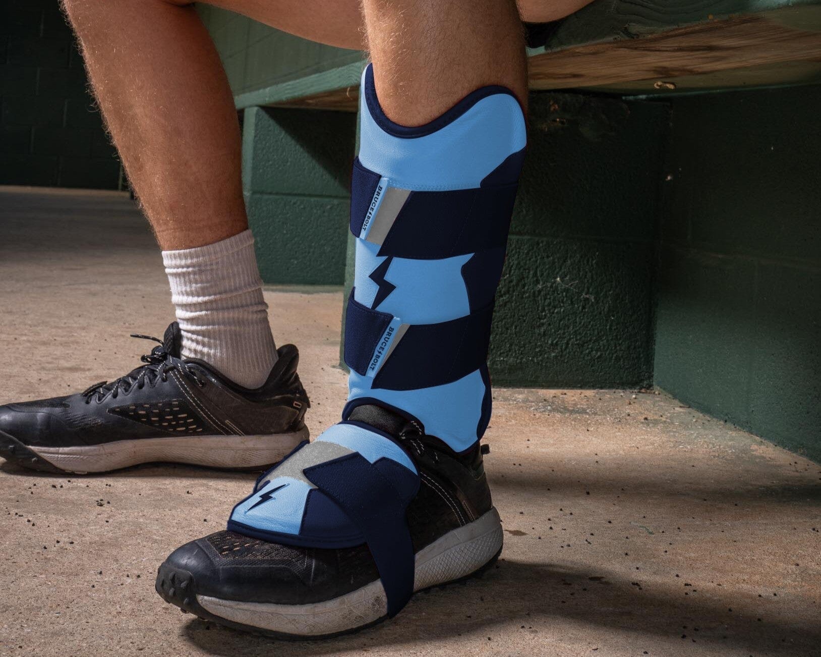 A close-up image of a person sitting down wearing a blue and dark blue BRUCE BOLT leg guard over their shoe.