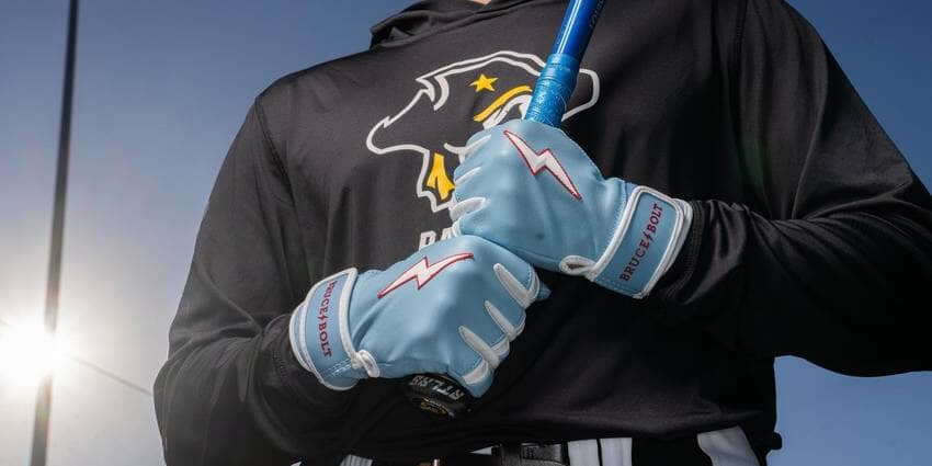 A close-up shot of a person wearing light blue BRUCE BOLT batting gloves with a red lightning bolt logo, holding a blue baseball bat.