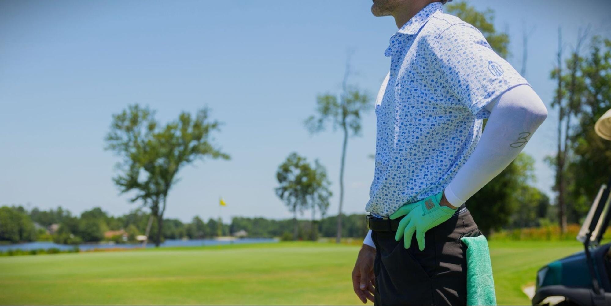 person wearing a white and blue patterned short-sleeve shirt, a light-colored arm sleeve, and a green golf glove stands on a sunny golf course with their hand on their hip. A golf cart and trees are visible in the background.
