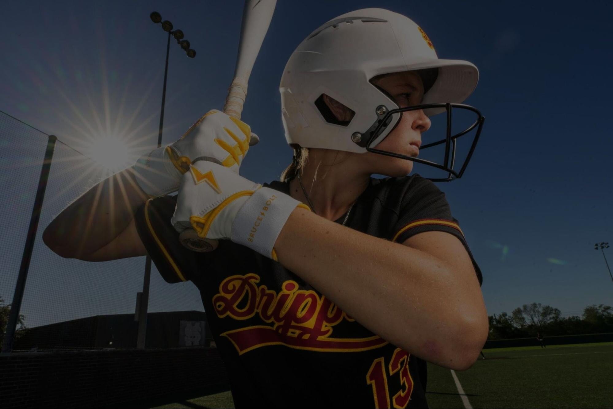 A close-up shot of a softball player in a batting stance, wearing a white helmet and white and yellow BRUCE BOLT batting gloves, holding a white bat, with the sun shining brightly in the background.